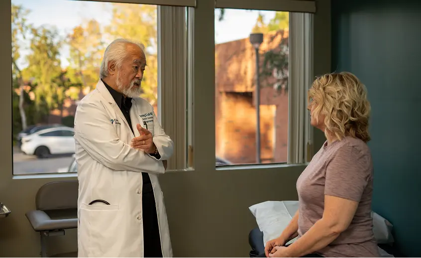 A healthcare worker speaks to a seated male patient in a medical clinic room with equipment and a white orchid on a side table.