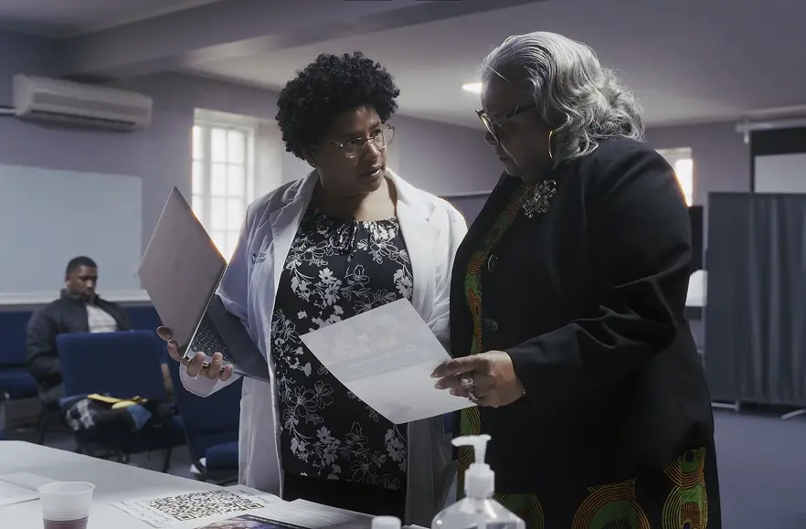 Two women stand at a table in an office, one holding a laptop and paper, the other holding a flyer. A man sits in the background. There are documents and sanitizer on the table.